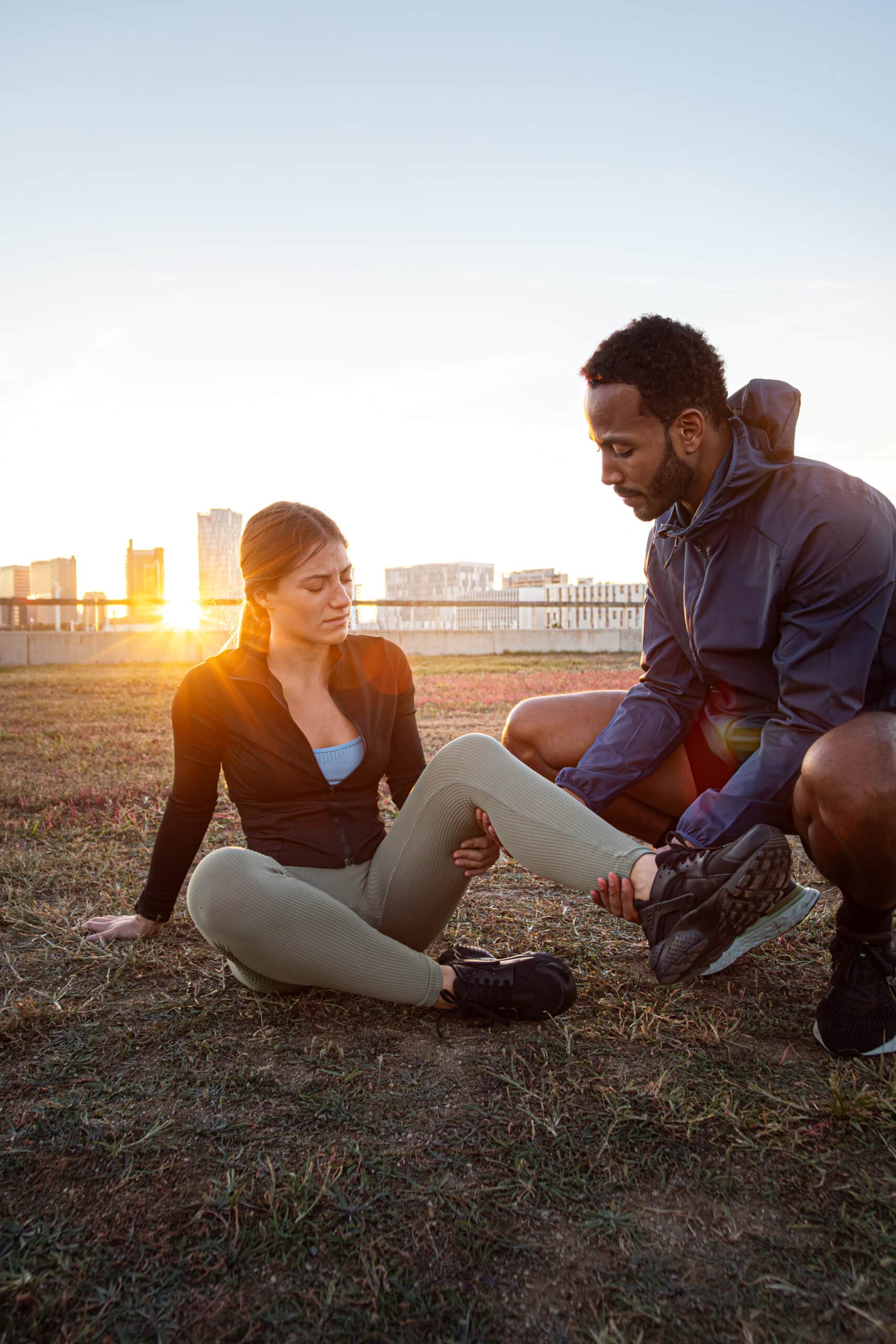 Personal trainer helping client with leg injury. Young woman sitting on the floor with knee pain during workout session. Vertical image. Copy space. Fitness and injury concept.