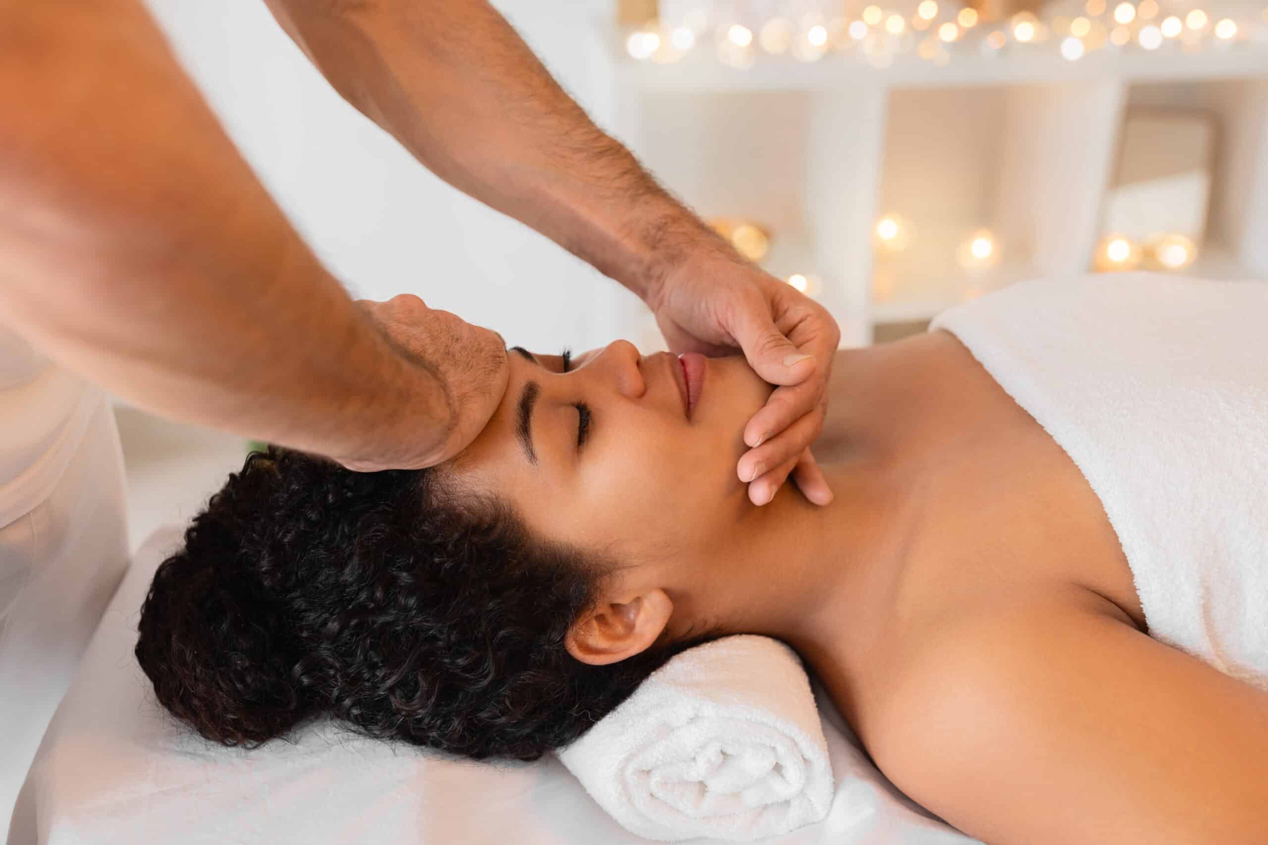 A skilled masseur provides a relaxing face massage to a client African American lady in a tranquil spa setting with dim lighting and candles